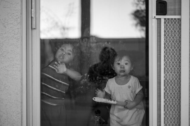 A grayscale photograph of a young child looking forlornly out of a window, in a sparsely furnished home, representing the economic strain on families due to the incarceration of a parent.