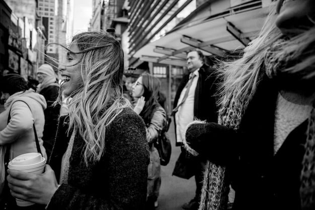 A black and white photograph depicting a long line of people waiting outside an immigration office in the United States. The faces show a mix of hope and weariness.
